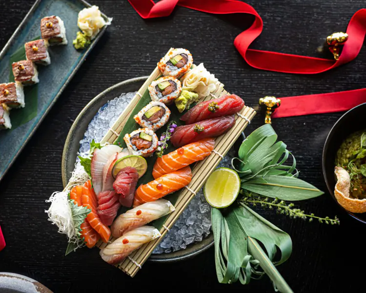 Assortment of sushi rolls on ice platter with red ribbon, garnishes, and black background