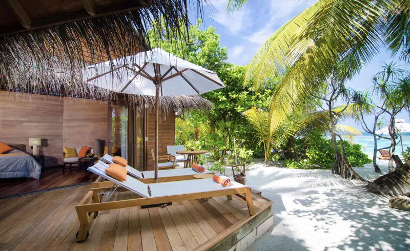 Luxury overwater villa at Mirihi Island Resort with lounge chairs on wooden deck, palm trees, white sand beach, and ocean.