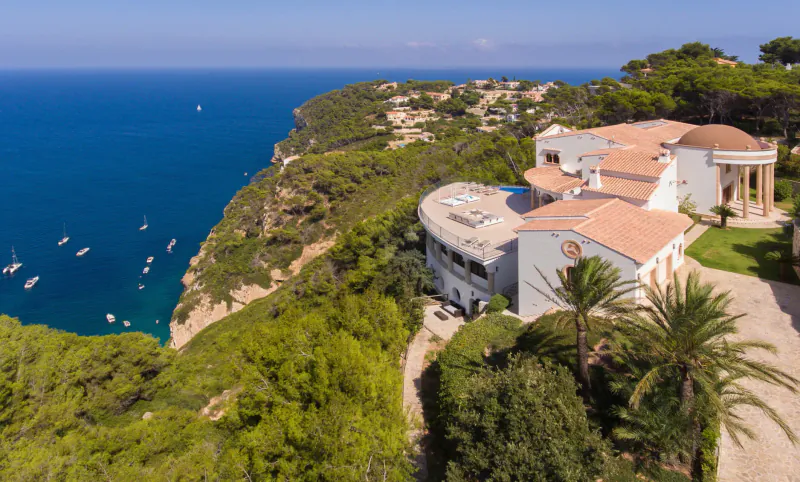 Aerial view of luxurious white villa with red-tiled roof on green cliff overlooking blue sea with yachts and pines.