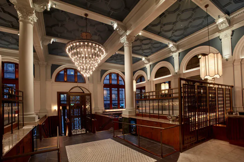 Elegant reimagined post office interior with grand chandelier, marble columns, and ornate wood bar in city centre gallery.