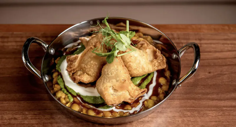 Four crispy samosas topped with greens in a silver balti bowl of chickpeas and sauce on wooden table
