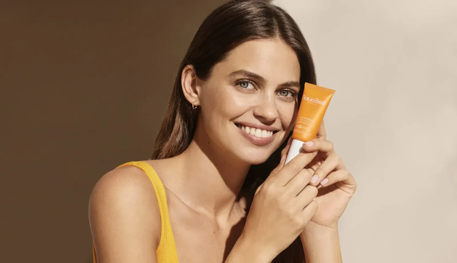 Smiling brunette woman in yellow top holding orange Natura Bissé skincare tube against warm backdrop