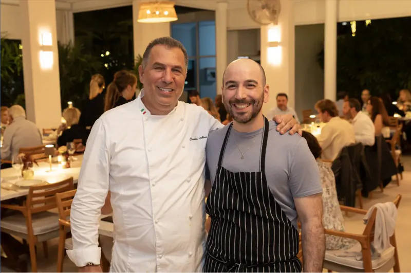Two smiling chefs in white uniform and apron pose arm-in-arm at outdoor restaurant dining area.