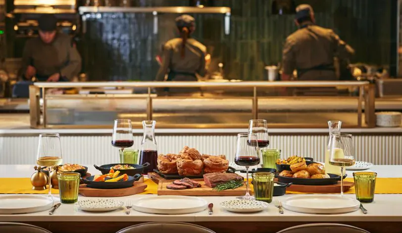 Park Corner Brasserie table with roast meats, Yorkshire puddings, vegetables, and wine glasses before blurred kitchen staff.