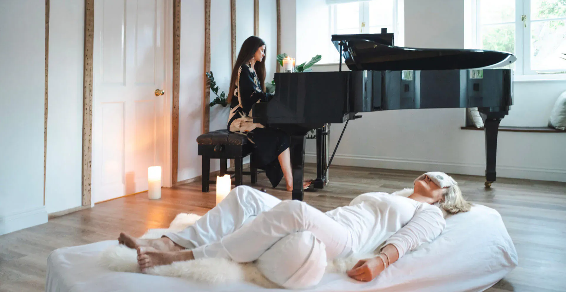 Woman in white lying relaxed on fur rug with eye mask; another at grand piano in serene room with candles.