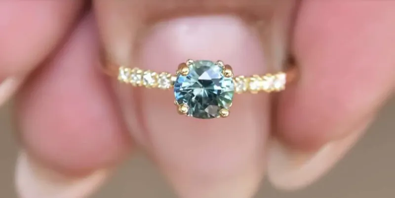 Close-up of gold engagement ring with blue-green gem and diamond side stones held in pink manicured fingers