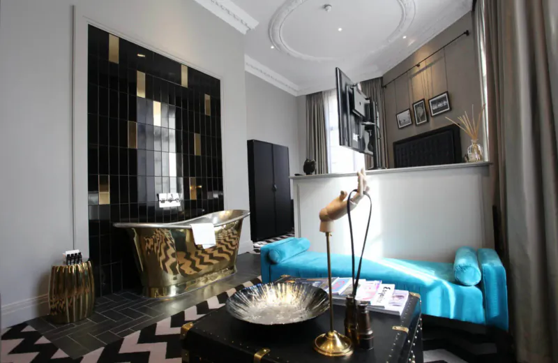 Luxurious Hotel Gotham bathroom with gold bathtub, turquoise chaise lounge, black-gold wall tiles, and modern decor.