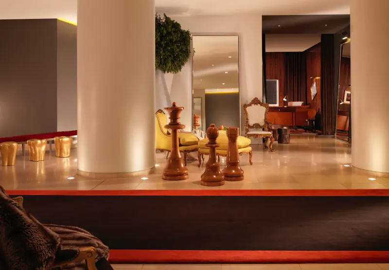 Elegant hotel lobby with large chess pieces on yellow chairs, potted plant, ornate furniture, columns, and warm lighting.