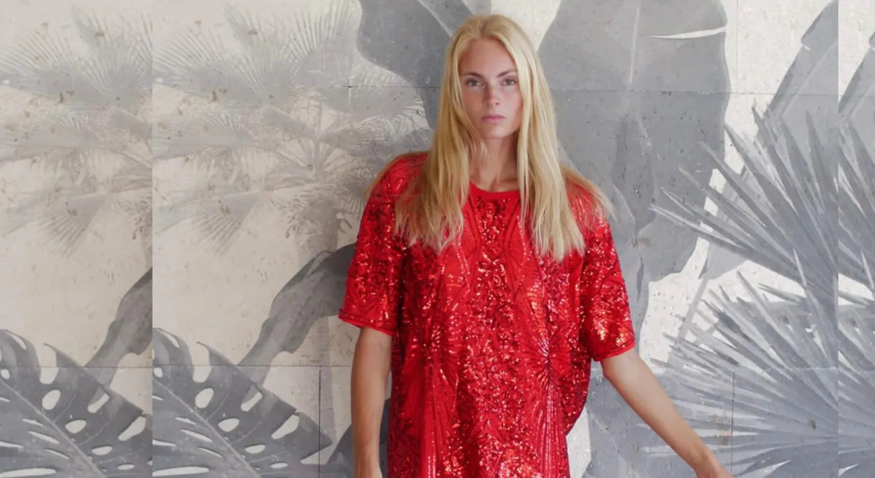 Blonde woman in sparkling red sequin dress poses against gray tropical leaf wall backdrop