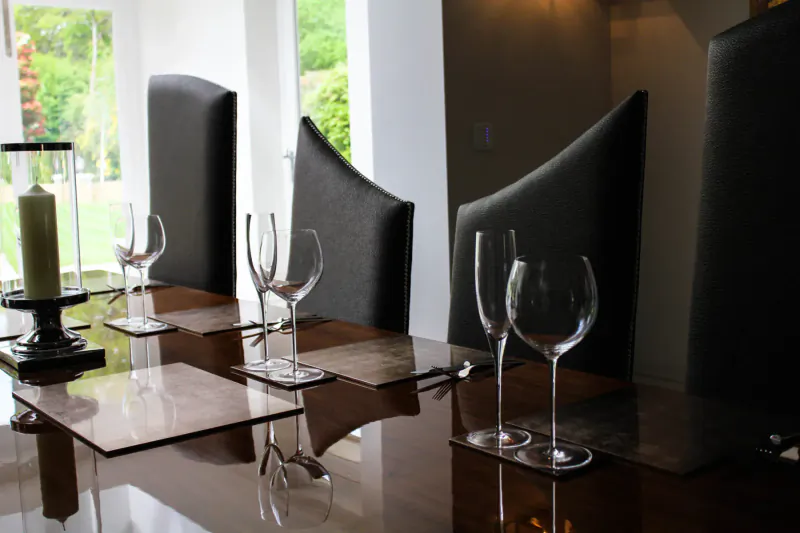 Elegant dining table set for two with wine glasses, placemats, candle, and curved black chairs by large window.