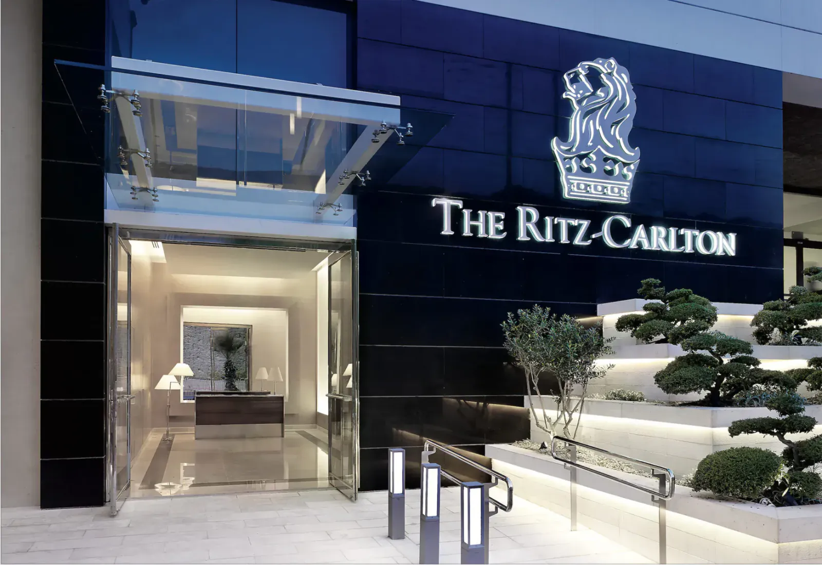 The Ritz-Carlton hotel entrance at night with lion crest logo, glass doors, lobby view, and bonsai plants.