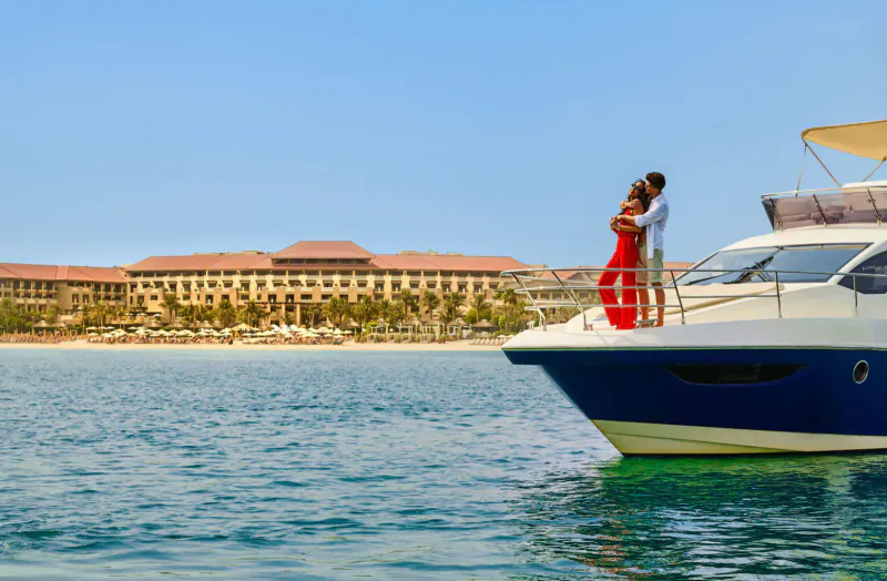 Couple embracing on luxury yacht railing, woman in red dress, Sofitel Dubai The Palm resort and sea in background.