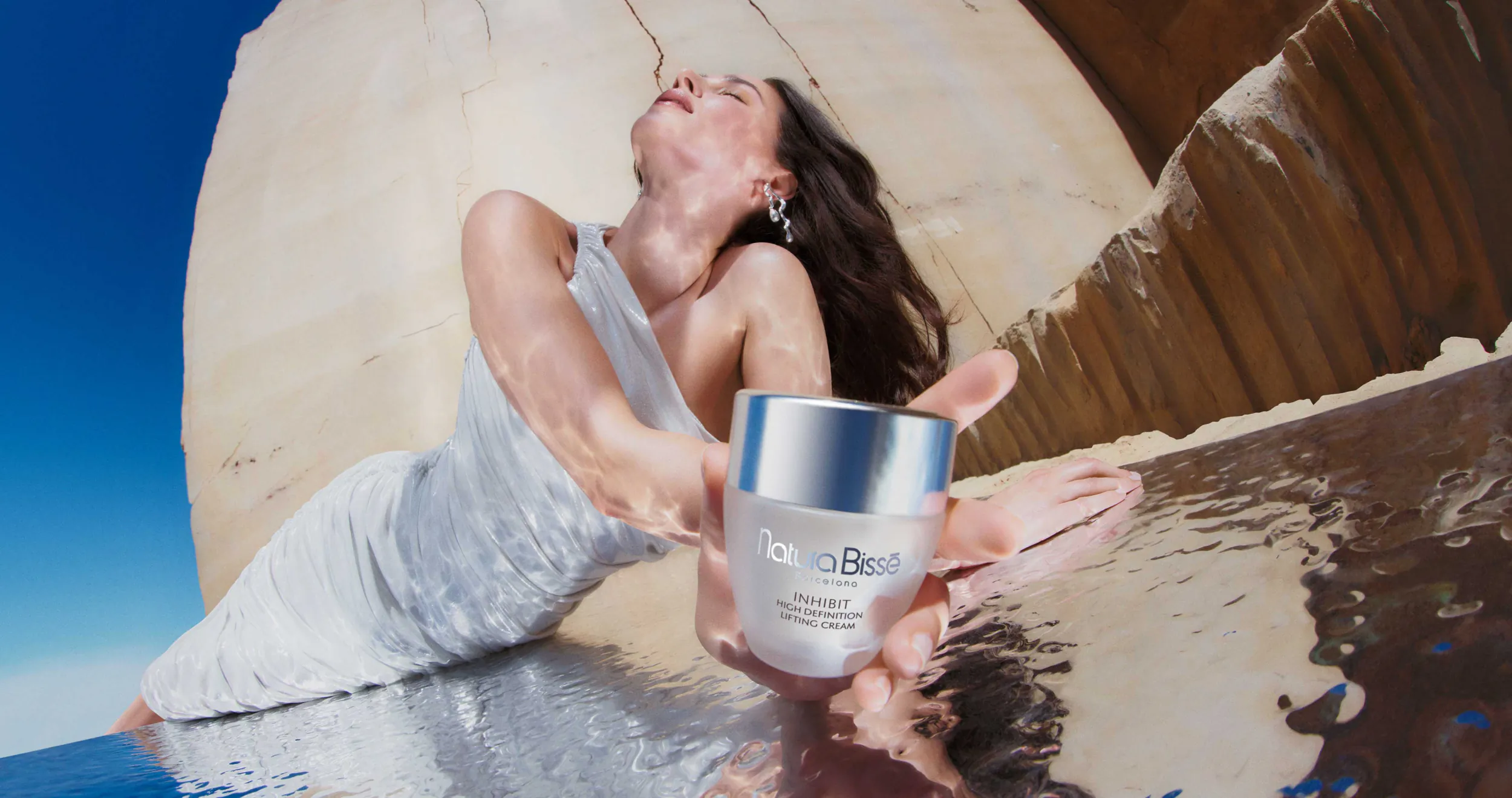 Woman in white dress reclining on beach, holding silver Beaute jar with water reflections and large rock backdrop