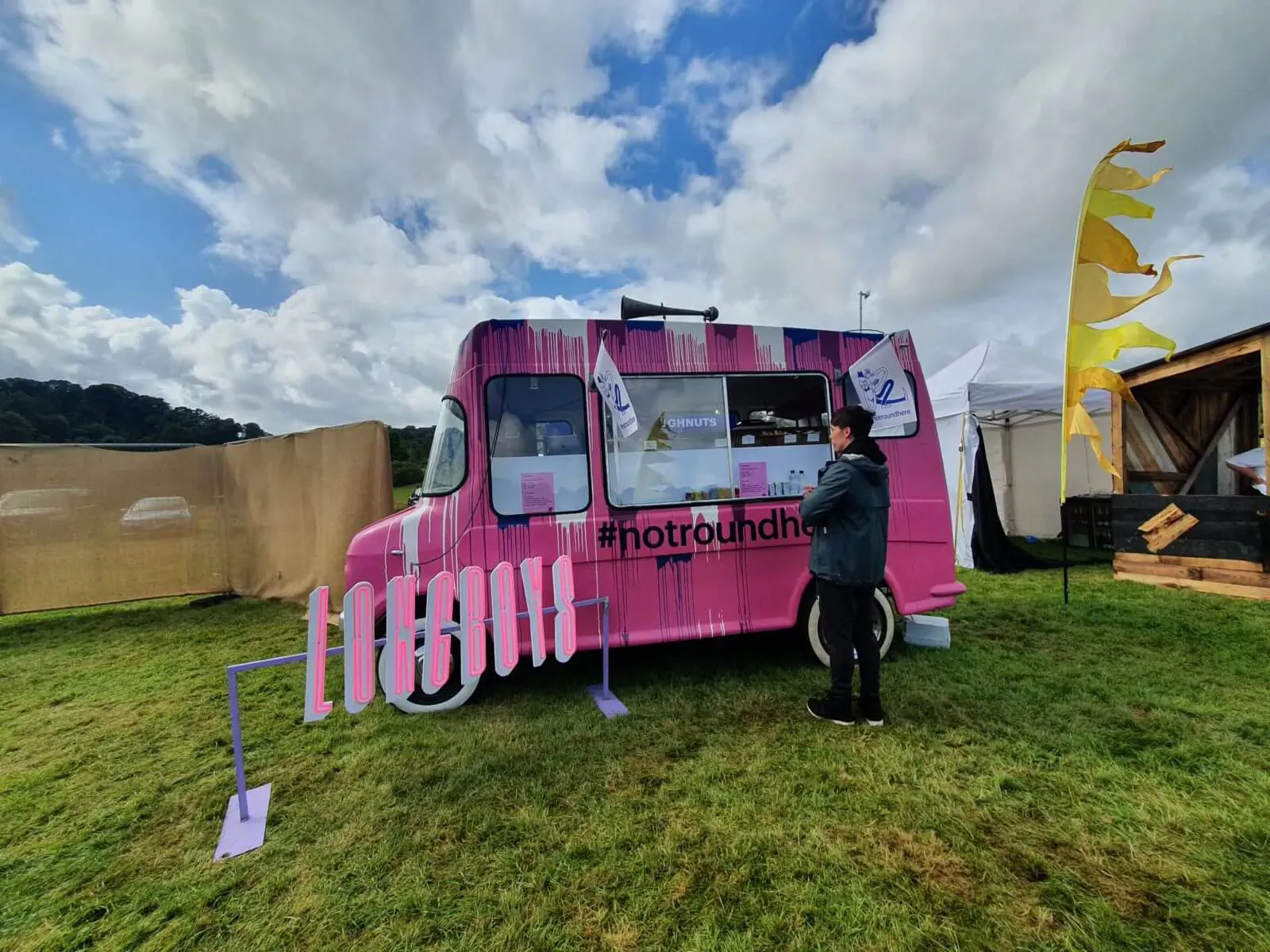 Pink #NotRound food truck at outdoor festival with Paul Ainsworth serving, cloudy sky, flags, and tents.