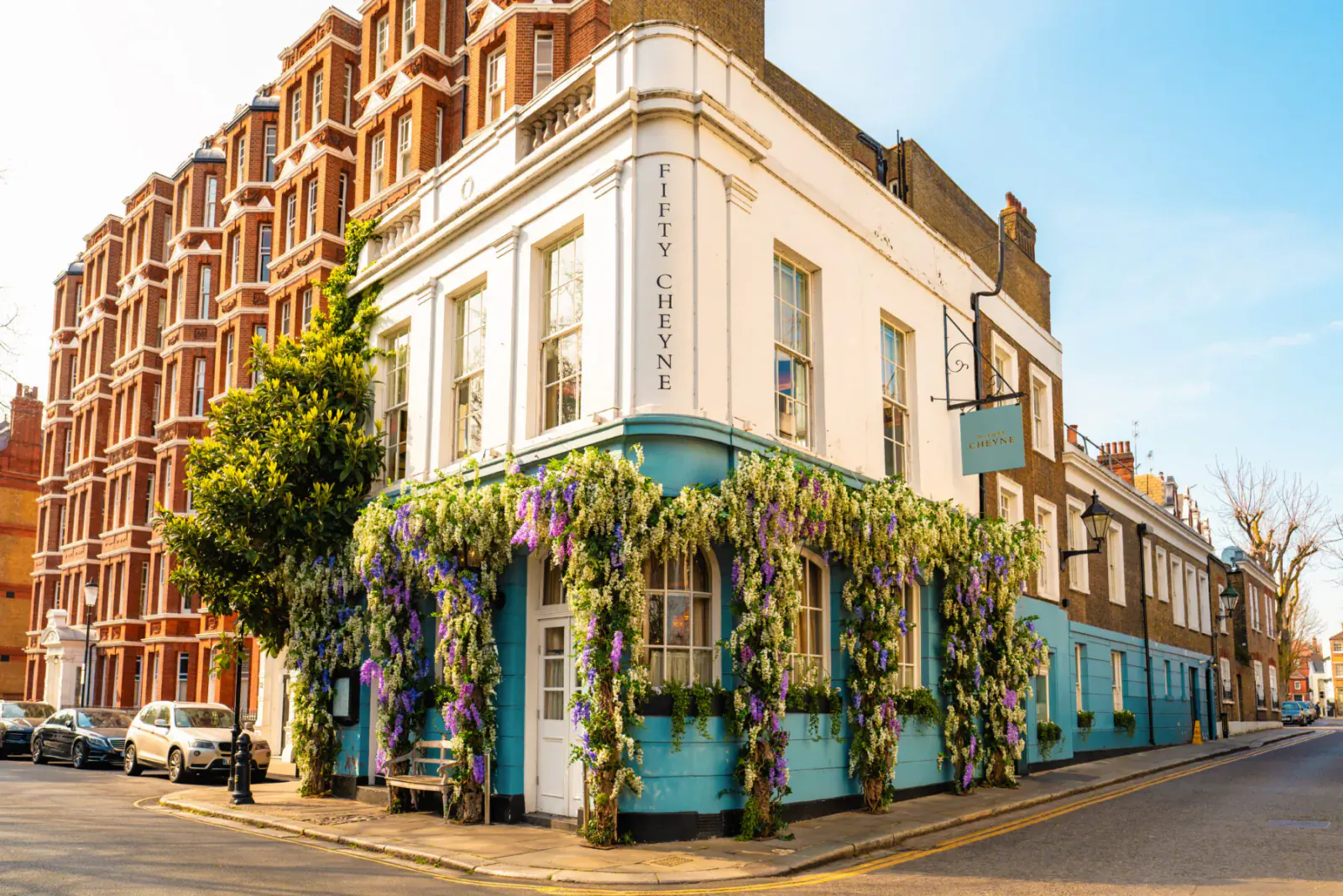 No. Fifty Cheyne pub on corner in Chelsea, white facade draped in purple flowers, red brick buildings, blue sky.