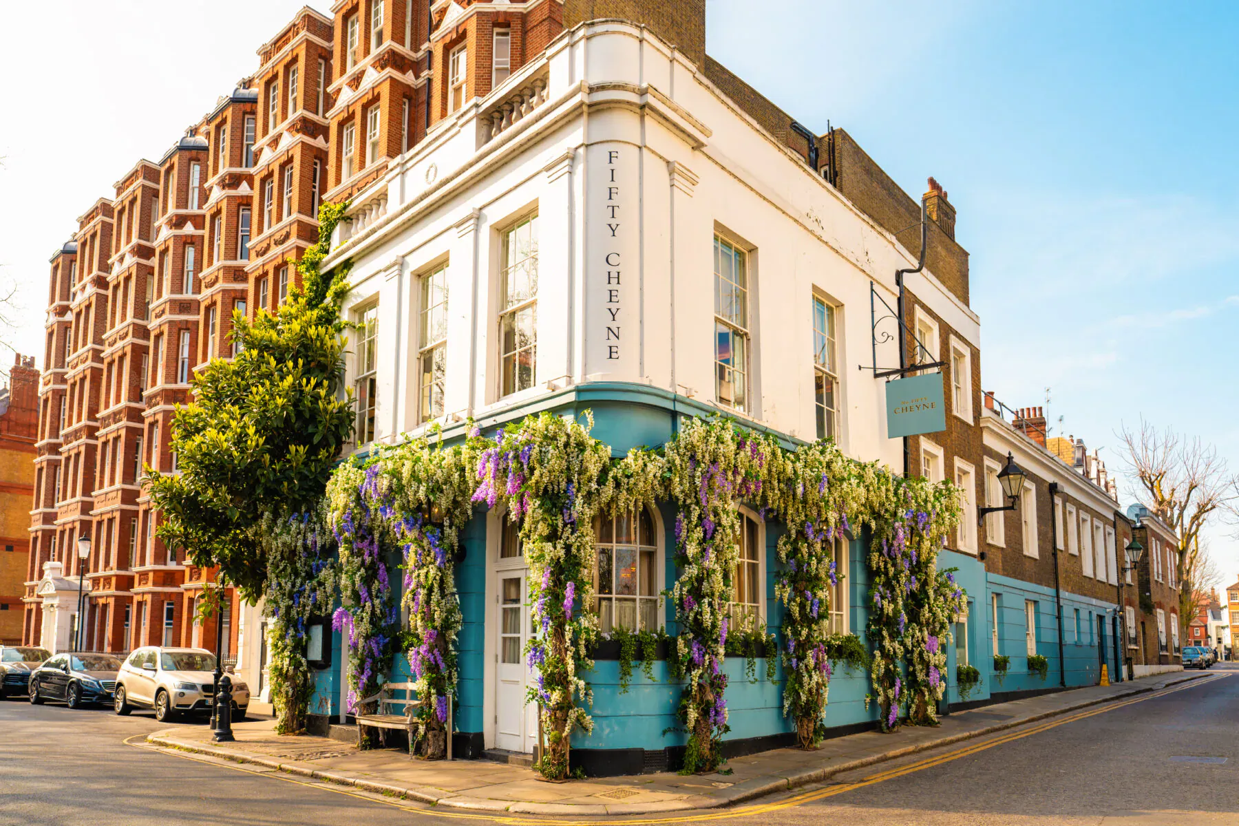 No. Fifty Cheyne pub on corner in Chelsea, white facade draped in purple flowers, red brick buildings, blue sky.