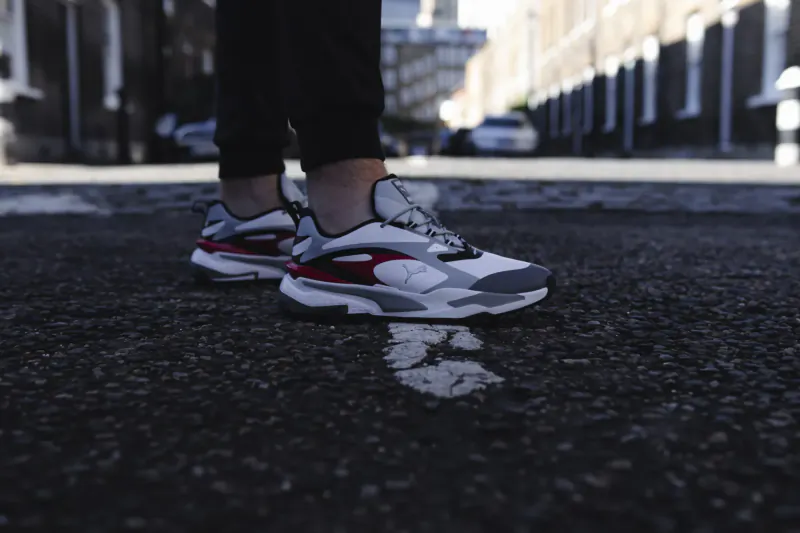 Close-up of white Puma golf sneakers with red accents on black pants, standing on urban street asphalt.