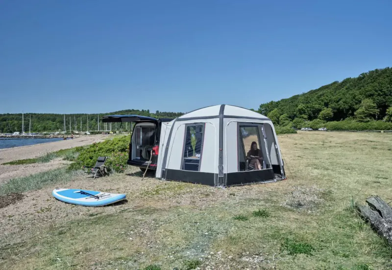 White Isabella motorhome awning pitched on grassy lakeside with hills, open door, person inside, blue paddleboard nearby.