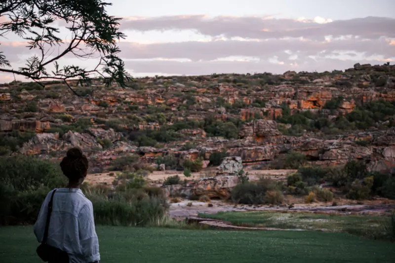 Woman with bun and backpack stands on green lawn facing layered red rock cliffs and river at Bushman’s Kloof sunset