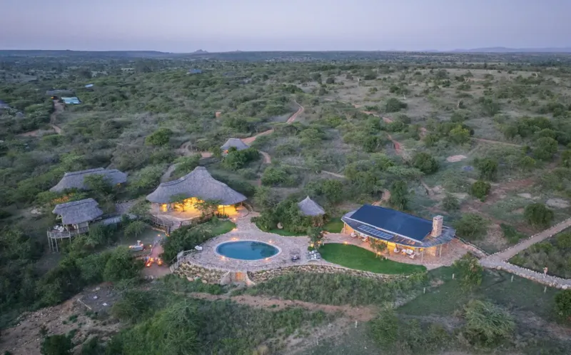 Aerial view of El Karama Lodge in Laikipia, Kenya: thatched huts, infinity pool, and solar-paneled building amid savanna at dusk.
