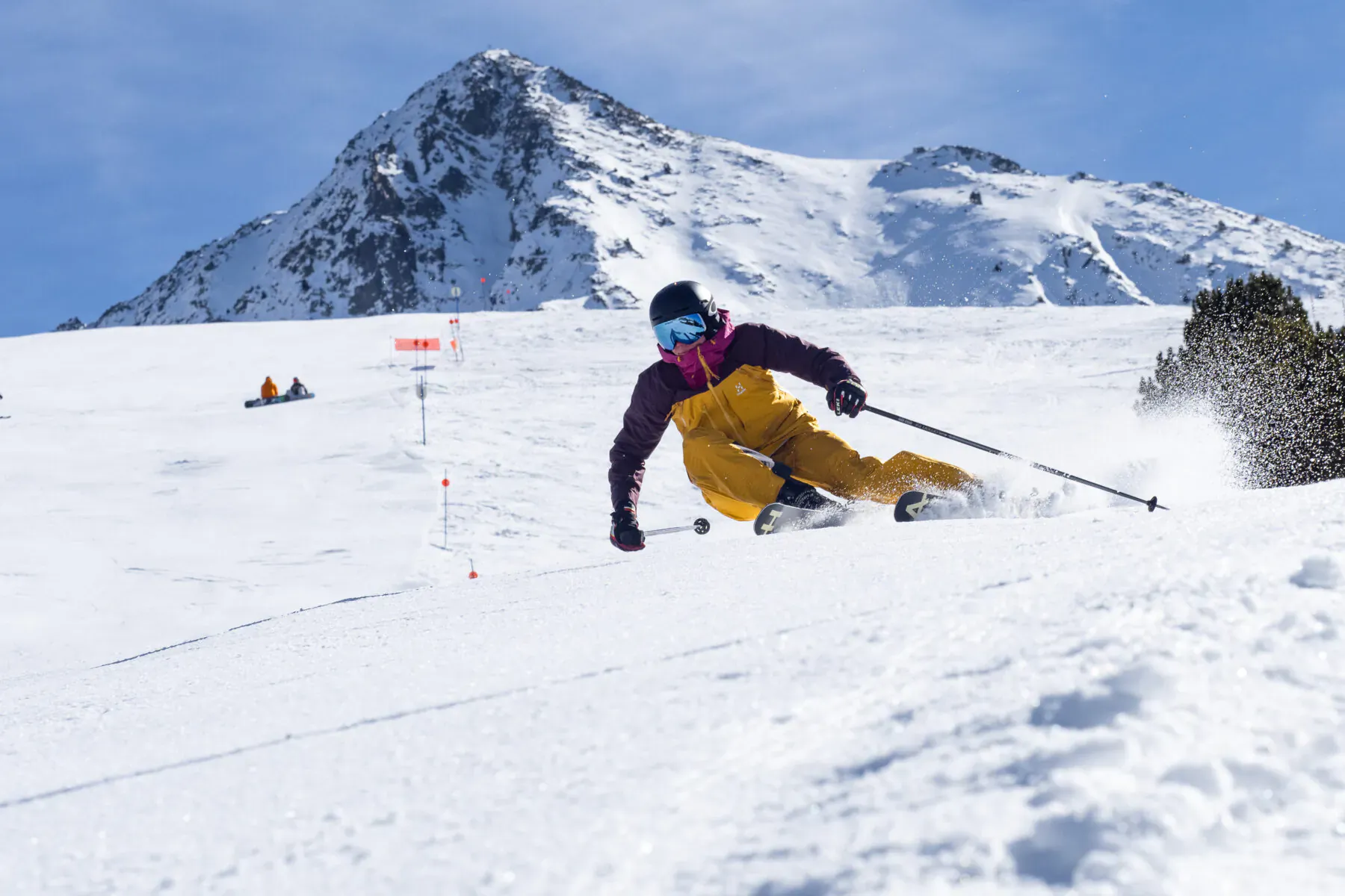 Skier in purple jacket and yellow pants carving down snowy Pyrenees slope in Val d'Aran, Catalonia, with mountain backdrop.