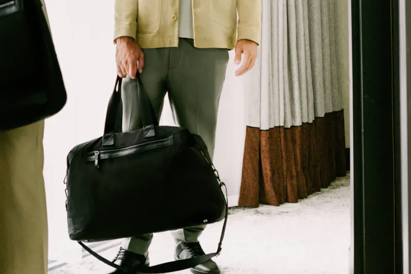 Man in beige jacket and gray pants holding black Antler Fenchurch duffel bag near hotel curtains