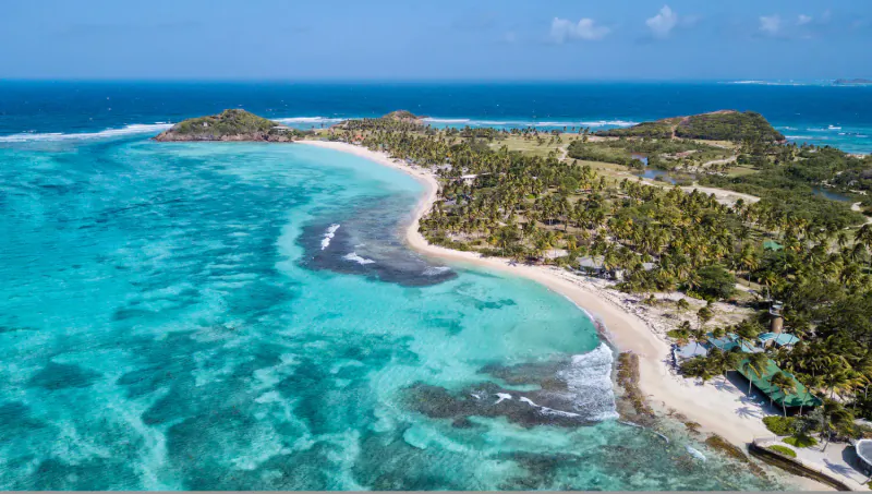 Aerial view of Palm Island Resort & Spa, curved white beach, turquoise waters, palm groves, and hills in St Vincent & Grenadines.