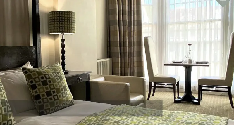 Elegant hotel room with green pillows on bed, plaid lamp, round table with glasses, armchairs, and large window.