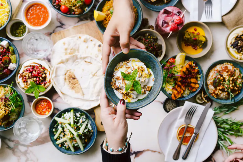 Hands passing flatbread over vibrant Bahraini dishes: dips, salads, veggies on wooden table