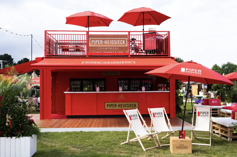 Piper-Heidsieck red Champagne Garden pavilion at Taste of London with bar, umbrellas, chairs, and plants on grass.