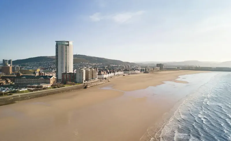 Aerial view of Swansea beach with Delta Hotel by Marriott, high-rise buildings, and hills under blue sky.