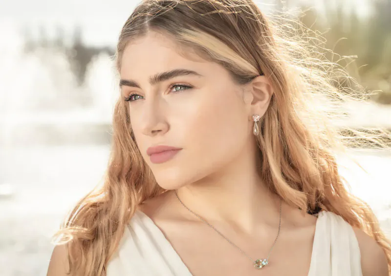 Young woman with wavy blonde hair and pearl earrings gazes sideways, wearing Augustine jewels necklace, Lake Como fountain backdrop
