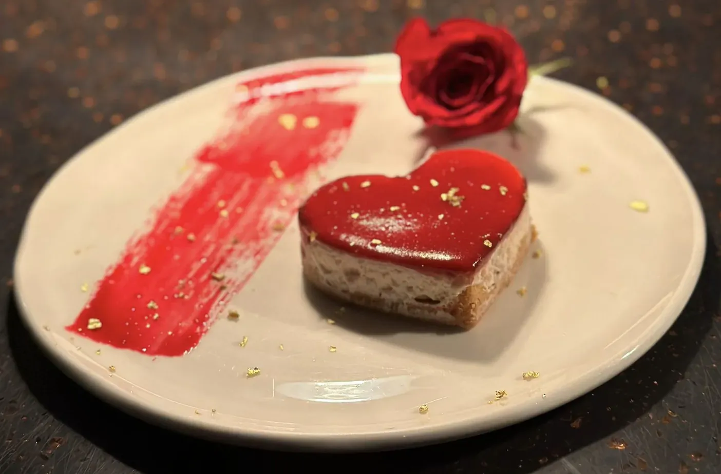 Heart-shaped red glazed cheesecake on white plate with red sauce streak, gold flecks, and red rose beside it.