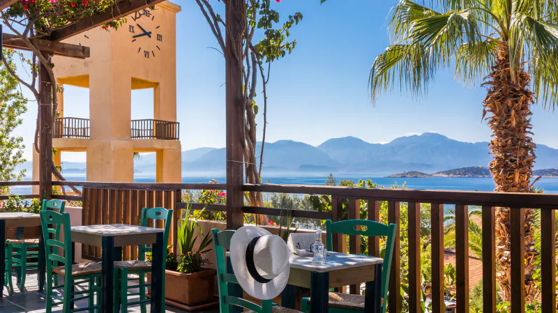 Outdoor terrace cafe at Candia Park Village, Crete, with green chairs, white tables, yellow clock tower, palm trees, and sea-mountain view.