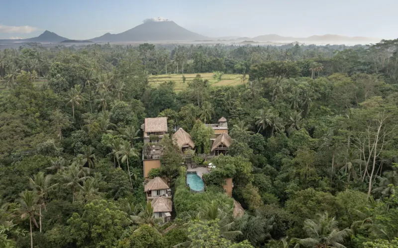 Aerial view of Kayon Resort Bali's thatched villas and pool nestled in lush jungle with volcano backdrop.