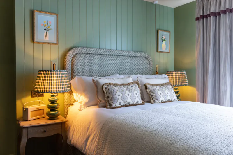 Cozy bedroom with white duvet on wicker headboard bed, green paneled walls, floral art, checkered lamps lit, in George & Dragon Marlow hotel.