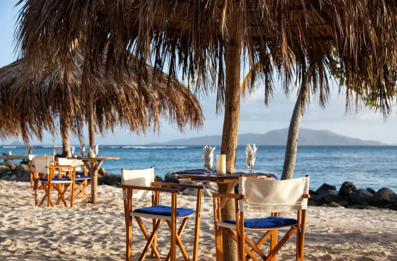 Beachfront dining setup at Palm Island Resort with thatched umbrellas, wooden tables, blue chairs, candles, and ocean view.