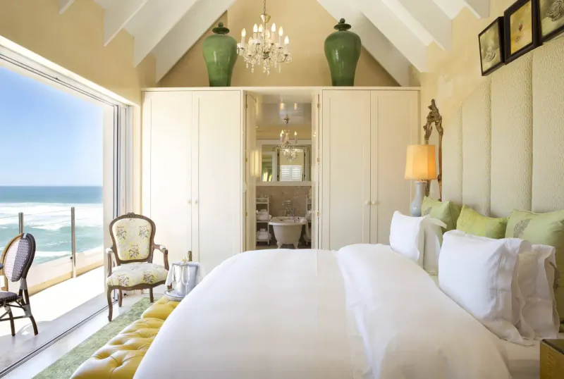 Luxurious bedroom in Hermanus with ocean view, white bed, green vases, chandelier, and en-suite bathroom.