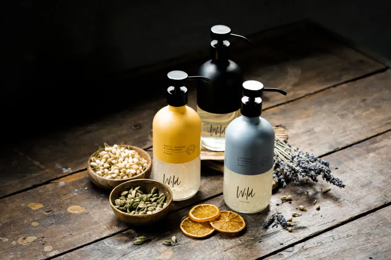 Three LUXE pump bottles in yellow, white, blue on wooden table with popcorn kernels, herbs, orange slices, lavender sprigs.