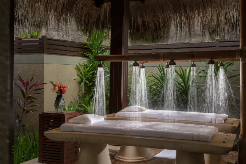 Outdoor tropical spa shower with water cascading over two white massage beds under thatched roof, surrounded by plants.