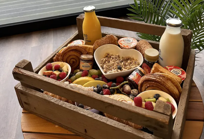 Wooden crate breakfast platter with oatmeal, croissants, fruits, yogurt, milk, and juice in glamping setting
