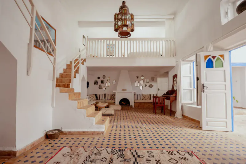 Spacious white Moroccan villa interior with wooden stairs, brass lantern, fireplace, zellige tiles, and blue doors