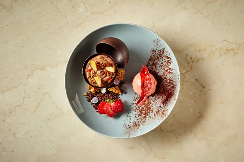 Kahani Indian dessert on blue plate: cracked chocolate egg with fruit and nuts, strawberry ice cream, fresh strawberries, cocoa dust.