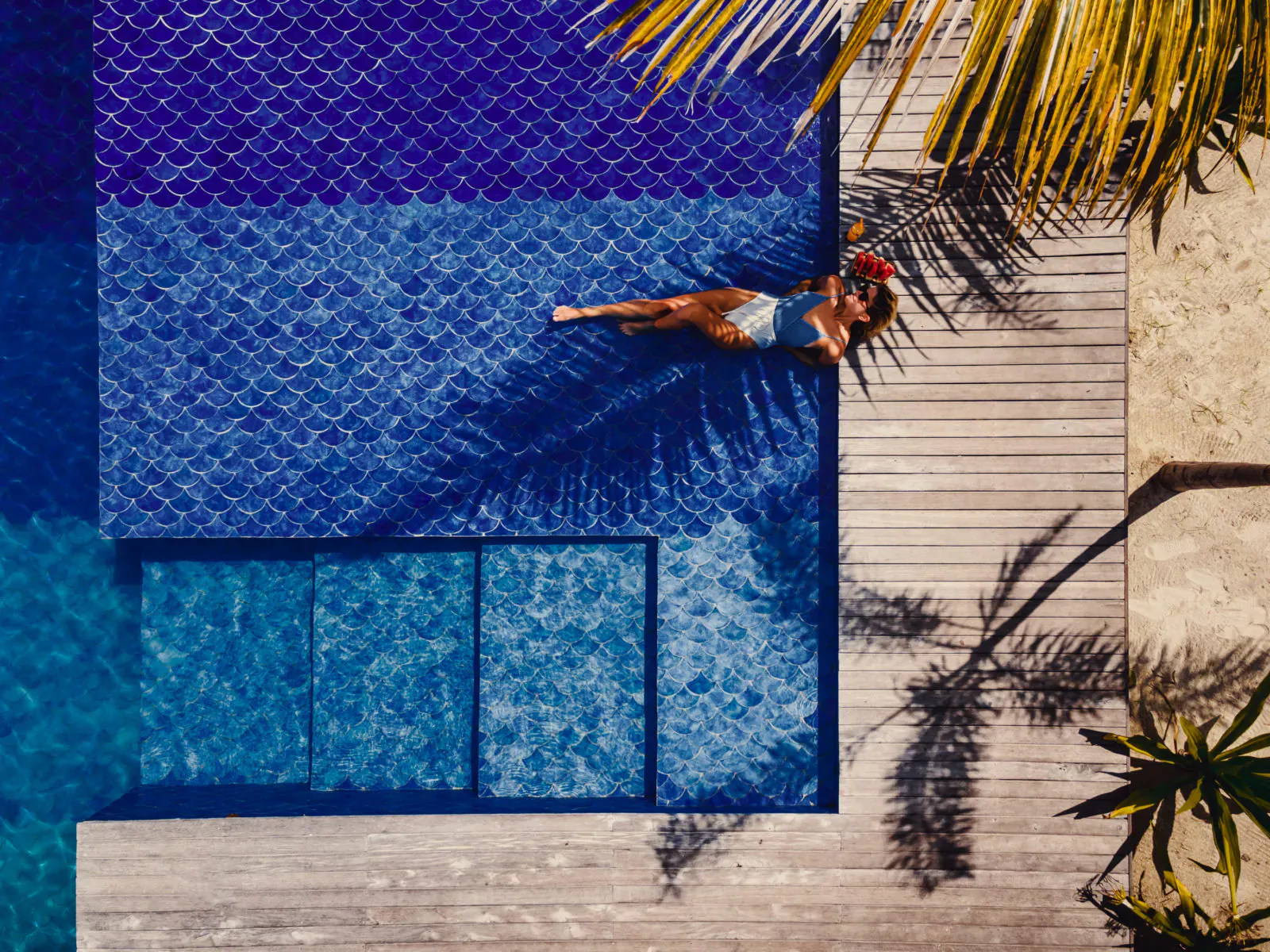 Aerial view of woman in blue swimsuit floating relaxed in mosaic infinity pool at beach resort, palm trees and wooden deck nearby.