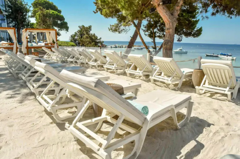 Rows of white lounge chairs with towels on sandy beach at Nikki Beach Ibiza, pine trees and sea view.