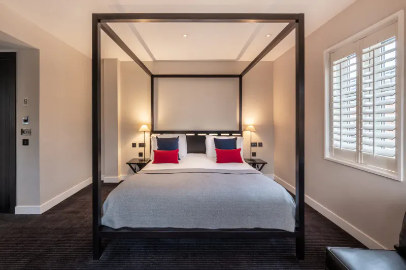Elegant four-poster bed with red pillows and gray bedding in a modern London hotel room with shutters.
