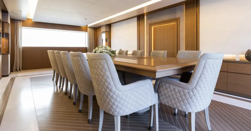 Elegant yacht dining room with long wooden table, white tufted chairs, wood paneling, and large windows.