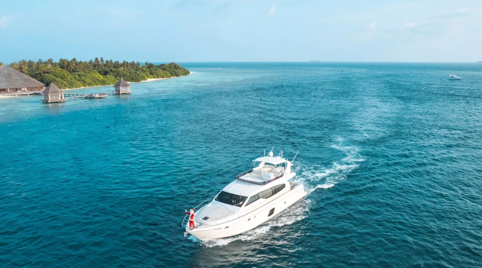 Aerial view of white luxury yacht Hideaway Majesty cruising in turquoise lagoon near tropical overwater villas