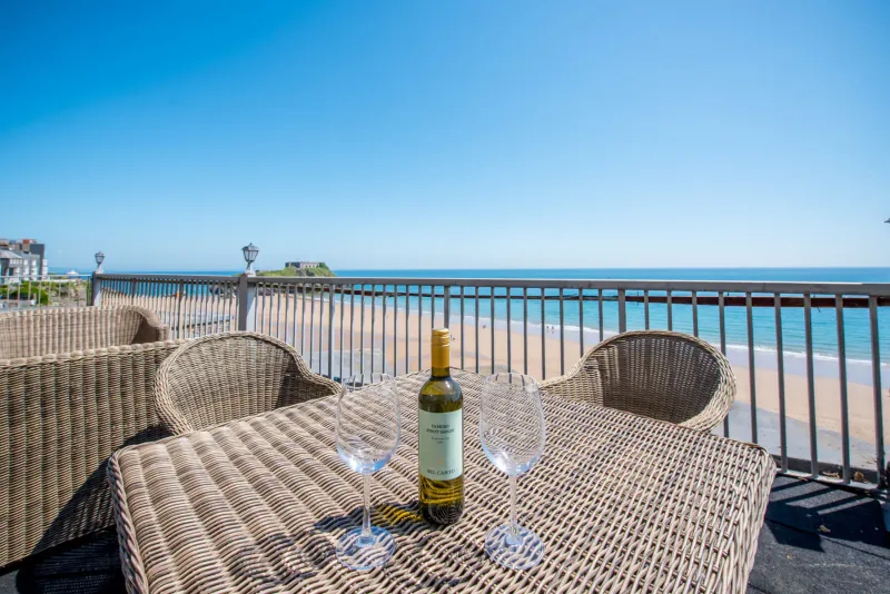 Wicker table on hotel balcony with wine bottle and glasses, overlooking sandy beach and ocean under blue sky.