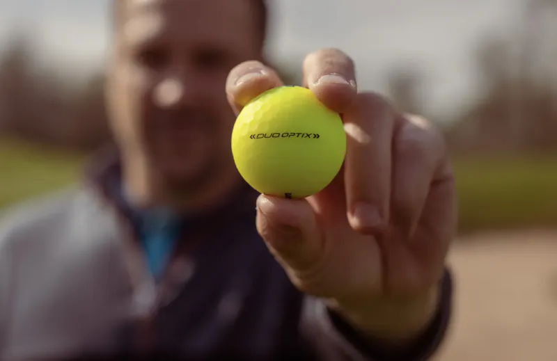 Man's hand holding yellow golf ball labeled 'Kulukopte' on golf course, Valentine's gift idea